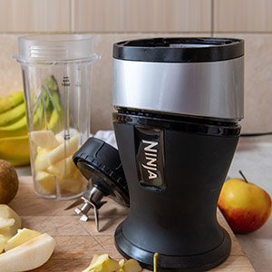 Black Ninja blender base, blade, and cup on a cutting board with fruit pieces.