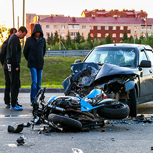 Two men stand next to a severe collision between a black car and a blue motorcycle in a residential area.